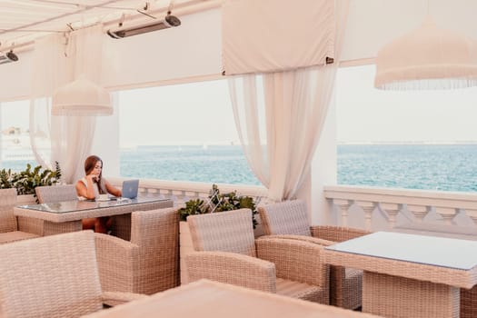Woman coffee cafe laptop sea. Female freelancer using laptop on coffee break in modern cafe with sea view in the morning.