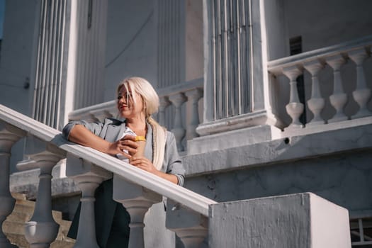 A woman is standing on a white railing with a cup of coffee in her hand. She is wearing a gray jacket and blue jeans. Concept of relaxation and leisure