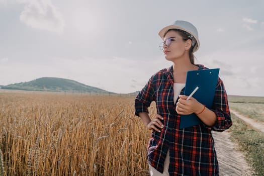 A young woman wearing a plaid shirt, white hardhat and glasses stands in a wheat field, holding a clipboard, pen, and looking to the horizon.