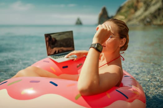Woman laptop sea. Freelancer woman in sunglases floating on an inflatable big pink donut with a laptop in the sea. People summer vacation rest lifestyle concept