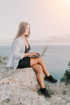 Digital nomad, woman in the hat, a business woman with a laptop sits on the rocks by the sea during sunset, makes a business transaction online from a distance. Freelance, remote work on vacation.