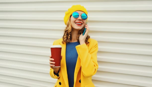 Autumn portrait of fashionable happy smiling young woman calling on phone, stylish girl talking on the smartphone holding cup of coffee in yellow french beret hat on white background