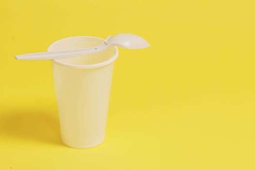 A white plastic glass and a spoon inserted into it on a yellow paper background.
