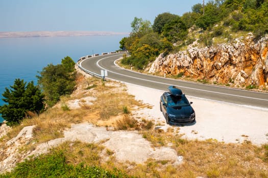 Black car with a roof box, parked at the edge of a narrow, winding road on a rugged cliff.