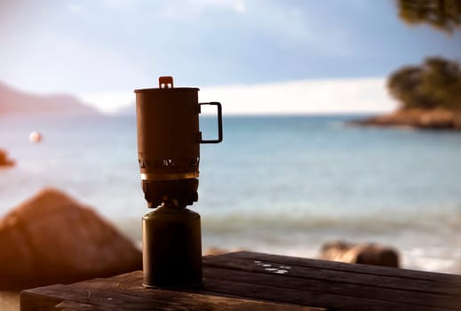 Camping burner stands on a table against the backdrop of a picturesque landscape with a beach and sea at sunset, camping equipment for quick cooking without the use of a fire with wood.