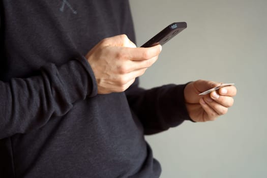 Male hands are holding a mobile phone and a bank card, a young man in a dark casual sweater pays for purchases, makes online banking using an application, close-up view.