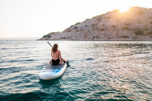 Woman paddle boarding on serene waters during sunset near a rocky shoreline.
