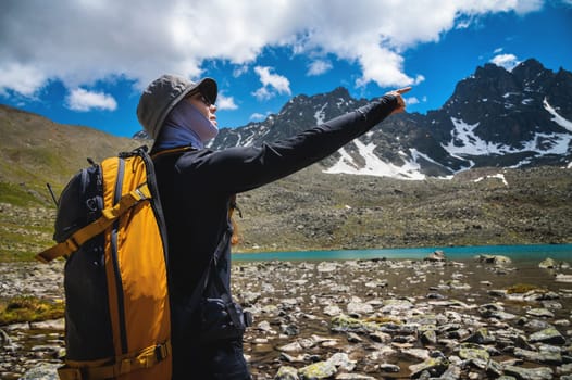 Close up of young Caucasian female traveler standing, showing destination mountains, pointing finger to right outdoors. Snowy mountains with a lake, a girl with a backpack on a sunny day.