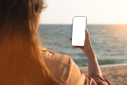 Hand holding smartphone on the beach, mobile blank screen mockup. Screen to place advertisements. Summer vacation concept, woman's hand with a phone on the background of the sea.