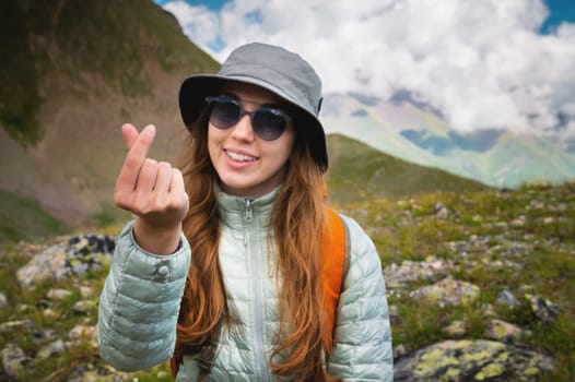 Attractive young Caucasian woman feels happy, shows a heart gesture, expresses tender feelings, against the backdrop of mountains and cloudy sky. Caring for people concept.