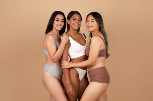 Positive young international women friends in different underwear, posing isolated on beige background, studio shot. Diversity, natural beauty care and fashion