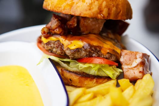 The concept of fast food and takeaway food. A juicy hamburger lies next to french fries on a metal plate along with cheddar cheese sauce in the background