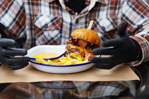The concept of fast food and takeaway food. A man in black latex gloves is ready to eat a juicy hamburger that lies near the french fries on a metal plate along with cheese sauce