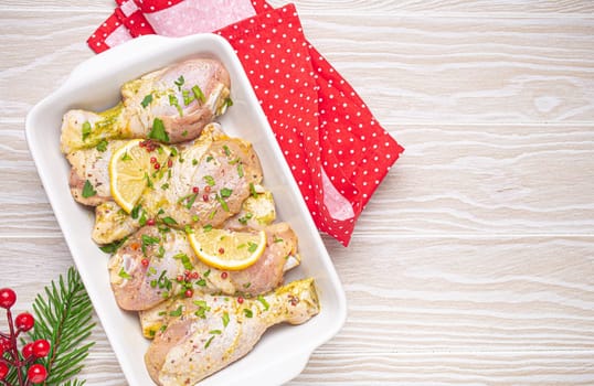 Preparing festive Christmas Eve meal with marinated chicken drumsticks. Raw chicken legs with herbs, lemon in white casserole top view on wooden kitchen table with Christmas decoration. Copy space