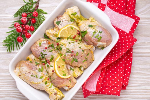 Preparing festive Christmas Eve meal with marinated chicken drumsticks. Raw chicken legs with herbs, lemon in white ceramic casserole top view on light wooden kitchen table with Christmas decoration.