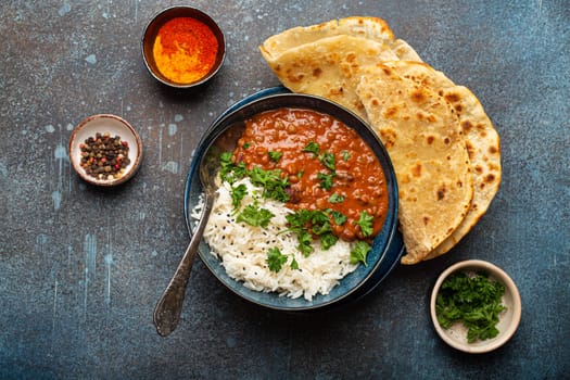 Traditional Indian Punjabi dish Dal makhani with lentils and beans in black bowl served with basmati rice, naan flat bread, fresh cilantro and spoon on blue concrete rustic table top view.