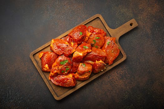 Raw uncooked chopped pieces of pork marinated with seasonings and parsley on wooden cutting board top view on dark rustic background. Cooking meal with marinated pork fillet pieces.