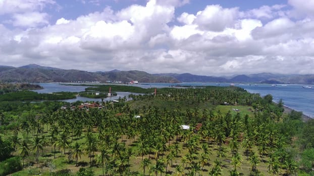 A breathtaking aerial view of Cemare Beach in Lombok, Indonesia, showcasing a lush coconut plantation, a tranquil bay, and majestic mountains under a dramatic sky.