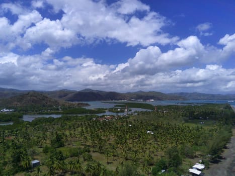 A breathtaking aerial view of Cemare Beach in Lombok, Indonesia, showcasing a lush coconut plantation, a tranquil bay, and majestic mountains under a dramatic sky.