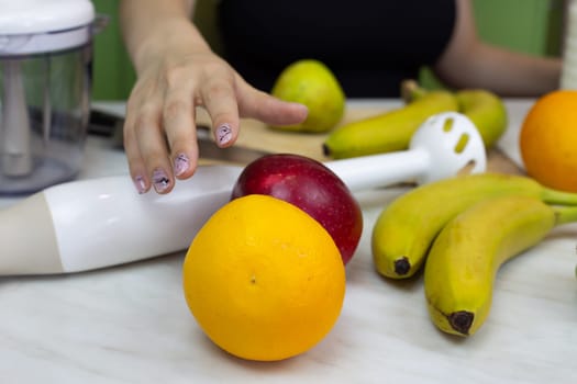 Girl reaches for an orange while preparing sports drink in the kitchen with blender. Girl makes healthy fruit cocktail to improve health, healthy food and lifestyle concept