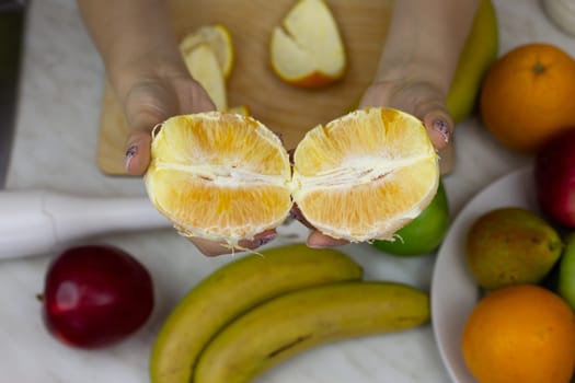 Two juicy halves of orange pulp in girl hands against the background of blender and fruits on table for sports nutrition. Vitamins and healthy lifestyle concept