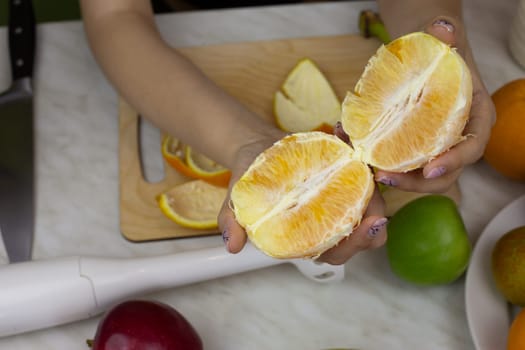 Peeled orange in girl hands against background of fruits on table. Healthy eating and lifestyle, making fruit drink to boost vitamins