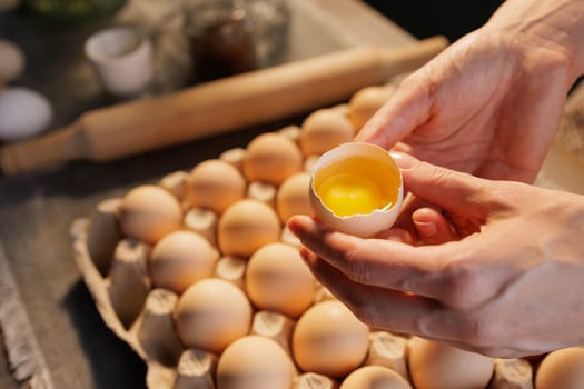 A person holding a cracked eggshell with a bright yellow yolk, surrounded by a tray of whole eggs. The setting is a kitchen with a rustic feel