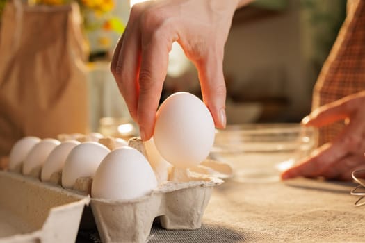 Hand taking chicken egg from box on kitchen table close-up against kitchen atmosphere