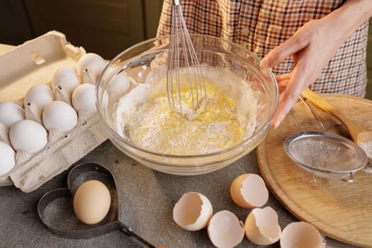 Close up of woman preparing dough in bowl against background of home kitchen, tray of eggs, eggshells and flour on table