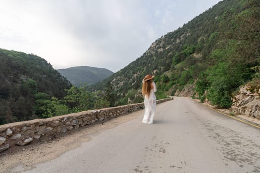 A woman in a white dress is walking down a road with a hat on. The road is surrounded by mountains and the sky is cloudy. The woman is enjoying the scenery and taking a break from her journey