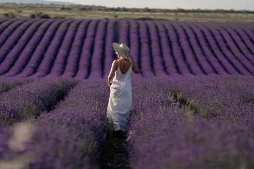 A woman is walking through a field of lavender. The field is vast and the flowers are purple. The woman is wearing a white dress and a straw hat