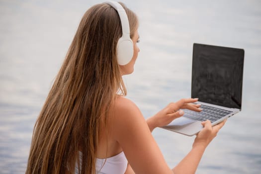 A woman is sitting on a beach with a laptop in her lap. She is wearing headphones and she is listening to music