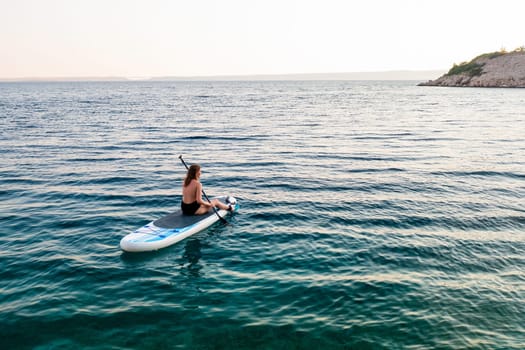 A woman paddle boarding on tranquil waters near a rocky shoreline at sunset.