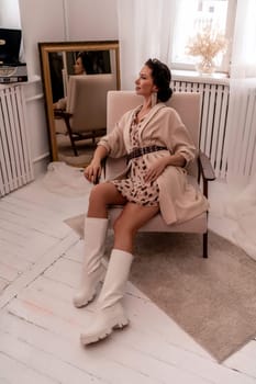 A woman is sitting in a chair wearing a white dress and white boots. She is wearing a brown jacket and a brown belt