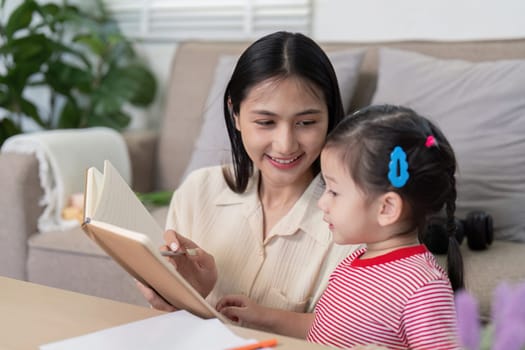 Asian mother guides her young daughter with a notebook, promoting learning and education in a cozy home environment.