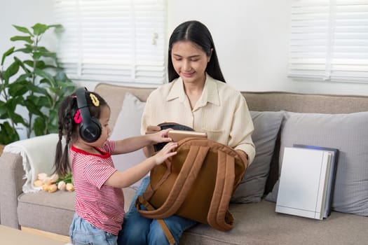 Little girl listens with headphones while her mother organizes school items in a home setting, highlighting back-to-school preparation