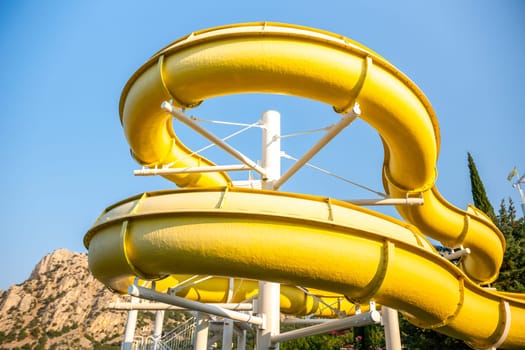 A yellow waterslide curves through the air against a bright blue sky. The slide is made of a smooth, shiny material and is supported by a white metal frame. family vacation concept