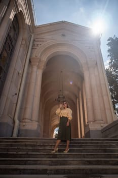 A woman in a skirt and sweater stands on steps leading up to a grand archway of an old building with pillars. The sun shines brightly in the sky.