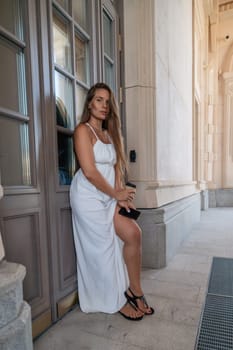 A woman in a white dress stands beside a wooden door, her right hand resting on the door frame. Her left hand holds a black purse. She is looking at the camera.
