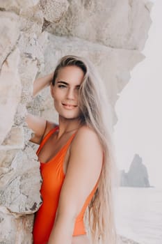 A young woman with long blonde hair is wearing an orange swimsuit and smiling while standing by rocks.