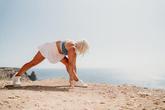 A blonde woman in athletic wear stretches her leg on a cliff overlooking the ocean, with a bright sky and sandy ground.