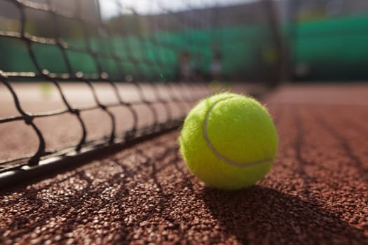 A close-up of a tennis ball resting on a clay court, with a net in the foreground and blurred players in the background