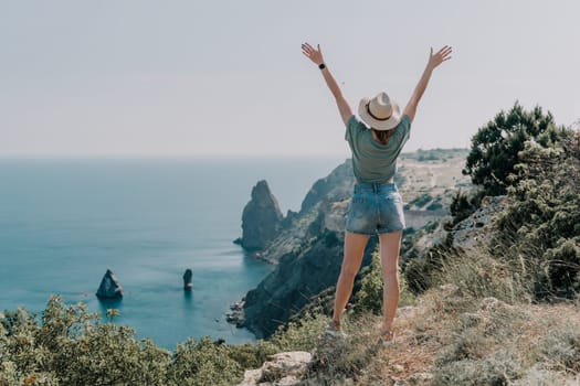 A woman in denim shorts and a hat stands with her arms raised in the air on a cliff overlooking the ocean.