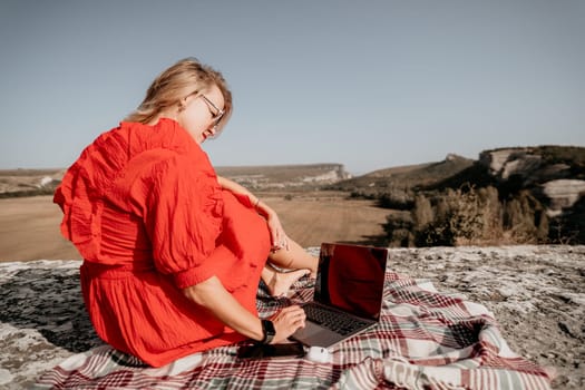 A woman in a red dress sits on a rock overlooking a valley, working on her laptop.