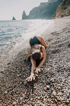 A woman in a black swimsuit reaches out with her hand towards the ocean waves on a beach covered in small stones.