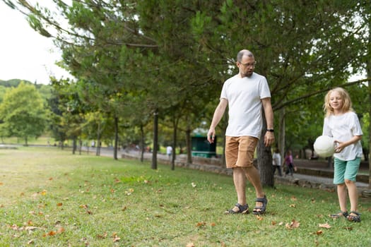 Father and daughter are enjoying a leisurely stroll through a lush green park, carrying a volleyball on a sunny day