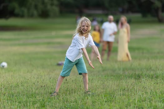 Young boy with long blond hair is playing in a park while his family walks in the background