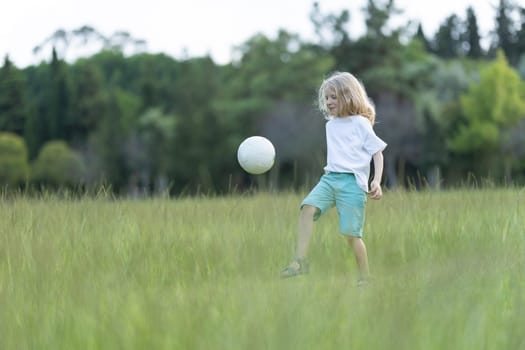 Young child is kicking a soccer ball in a field with tall grass and trees in the background