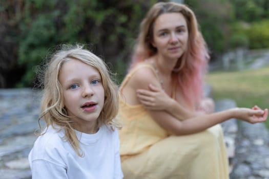 Young blonde girl with blue eyes looking pensive while sitting with her mother outdoors