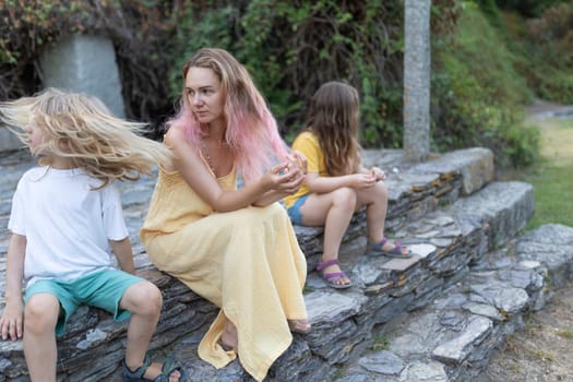 Mother with pink hair wearing a yellow dress is sitting on stone steps with her two daughters, seemingly sad and thoughtful